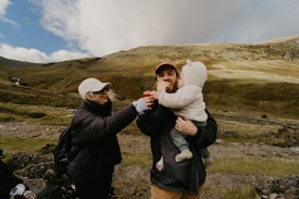A family scene in a mountainous landscape, with grass-covered hills under a partly cloudy sky. An adult woman wearing sunglasses and a hat is playfully feeding a man holding a toddler in a fuzzy outfit.