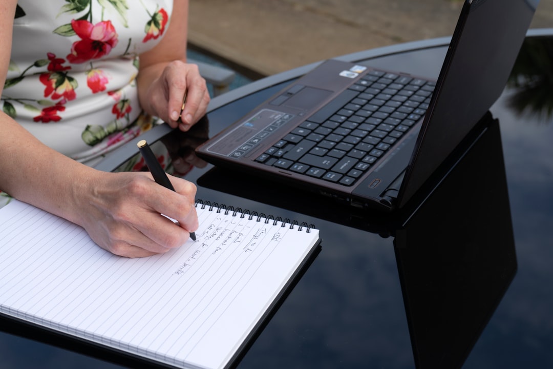 Woman writing in a notebook and using a laptop computer whilst working from home outside in the garden