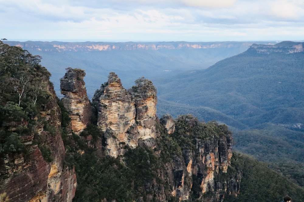 a rocky cliff with trees and a valley below with Three Sisters in the background