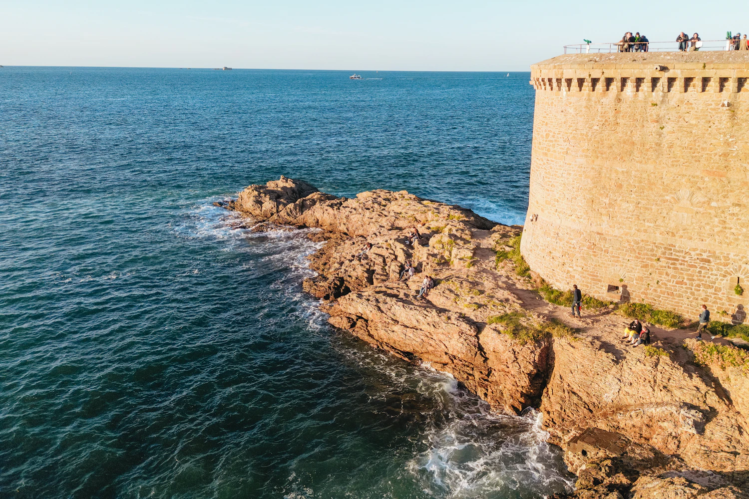 A round stone bastion standing on a rocky headland above the sea, people walking along the top of the wall