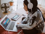A person wearing a patterned sweater and headphones is using a laptop, likely engaged in music production. A MIDI keyboard is nearby, and a fan sits on a table in the background near a potted plant.