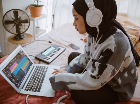 A person wearing a patterned sweater and headphones is using a laptop, likely engaged in music production. A MIDI keyboard is nearby, and a fan sits on a table in the background near a potted plant.