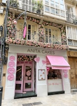 A charming and inviting bakery storefront decorated with numerous pink and white flowers around the entrance and windows. The signage reads 'ANITA CAKES' in simple, elegant letters above the doorway. Heart-shaped signs with messages like 'LOVE', 'BE SWEET', and 'TAKE ME AWAY' are displayed outside, enhancing the shop's playful and romantic theme. A pink awning with the bakery's logo adds to the visual appeal.