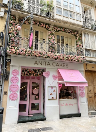 A charming and inviting bakery storefront decorated with numerous pink and white flowers around the entrance and windows. The signage reads 'ANITA CAKES' in simple, elegant letters above the doorway. Heart-shaped signs with messages like 'LOVE', 'BE SWEET', and 'TAKE ME AWAY' are displayed outside, enhancing the shop's playful and romantic theme. A pink awning with the bakery's logo adds to the visual appeal.