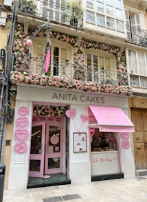A charming and inviting bakery storefront decorated with numerous pink and white flowers around the entrance and windows. The signage reads 'ANITA CAKES' in simple, elegant letters above the doorway. Heart-shaped signs with messages like 'LOVE', 'BE SWEET', and 'TAKE ME AWAY' are displayed outside, enhancing the shop's playful and romantic theme. A pink awning with the bakery's logo adds to the visual appeal.