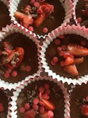 Several chocolate cupcake batter portions are placed in white and pink paper liners, topped with slices of strawberries, pink wafer pieces, and chocolate shavings.
