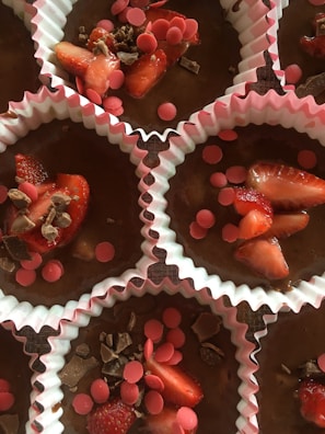 Several chocolate cupcake batter portions are placed in white and pink paper liners, topped with slices of strawberries, pink wafer pieces, and chocolate shavings.