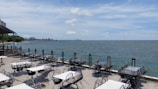 A seaside outdoor dining area featuring numerous empty tables and chairs arranged on a patio overlooking the ocean. The sky is partly cloudy with patches of blue, and the water extends to the horizon. In the distance, a coastline with buildings and greenery is visible.