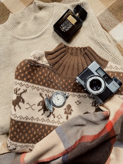 Several cozy items arranged on a plaid blanket include two knit sweaters, one with a reindeer pattern and one plain, a vintage camera, a pocket watch with a chain, and a dark bottle of black perfume.