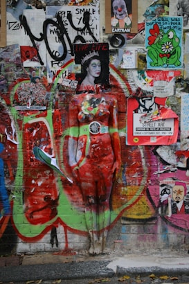 A vibrant collage of street art on a wall, featuring a mix of posters, graffiti, and a painted mannequin. The mannequin is painted to blend with the background, with bright colors and patterns. Various stickers and posters include phrases and artistic designs, contributing to a chaotic yet creative atmosphere.