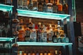 A variety of liquor bottles displayed on illuminated shelves against a brick wall. The setup includes different brands and types of alcohol, with glassware hanging nearby. The shelves are highlighted with a blue-green light.