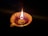 A decorative clay lamp with a visible wick burns brightly, casting a warm glow. The lamp is designed with a floral petal shape and is set against a dark background that enhances its illumination.