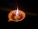 A decorative clay lamp with a visible wick burns brightly, casting a warm glow. The lamp is designed with a floral petal shape and is set against a dark background that enhances its illumination.