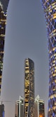 A modern cityscape displaying tall, uniquely designed skyscrapers. The central building features a sleek, curved design and is surrounded by other illuminated structures with geometric patterns and blue lighting.