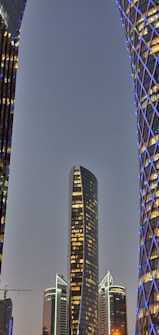 A modern cityscape displaying tall, uniquely designed skyscrapers. The central building features a sleek, curved design and is surrounded by other illuminated structures with geometric patterns and blue lighting.