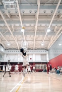 A volleyball player mid-jump, preparing to spike the ball in a bright indoor court. Other players are visible in the background, wearing matching uniforms and observing the play. The gymnasium features a high ceiling with visible structural beams and lighting fixtures, and there are spectators seated on benches to the side.
