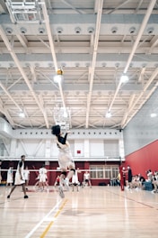 A volleyball player mid-jump, preparing to spike the ball in a bright indoor court. Other players are visible in the background, wearing matching uniforms and observing the play. The gymnasium features a high ceiling with visible structural beams and lighting fixtures, and there are spectators seated on benches to the side.