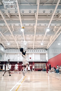 A volleyball player mid-jump, preparing to spike the ball in a bright indoor court. Other players are visible in the background, wearing matching uniforms and observing the play. The gymnasium features a high ceiling with visible structural beams and lighting fixtures, and there are spectators seated on benches to the side.