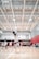 A volleyball player mid-jump, preparing to spike the ball in a bright indoor court. Other players are visible in the background, wearing matching uniforms and observing the play. The gymnasium features a high ceiling with visible structural beams and lighting fixtures, and there are spectators seated on benches to the side.