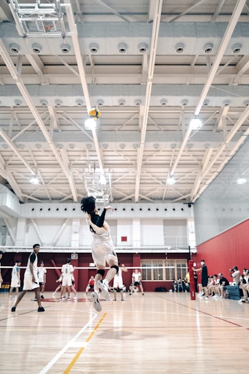 A volleyball player mid-jump, preparing to spike the ball in a bright indoor court. Other players are visible in the background, wearing matching uniforms and observing the play. The gymnasium features a high ceiling with visible structural beams and lighting fixtures, and there are spectators seated on benches to the side.