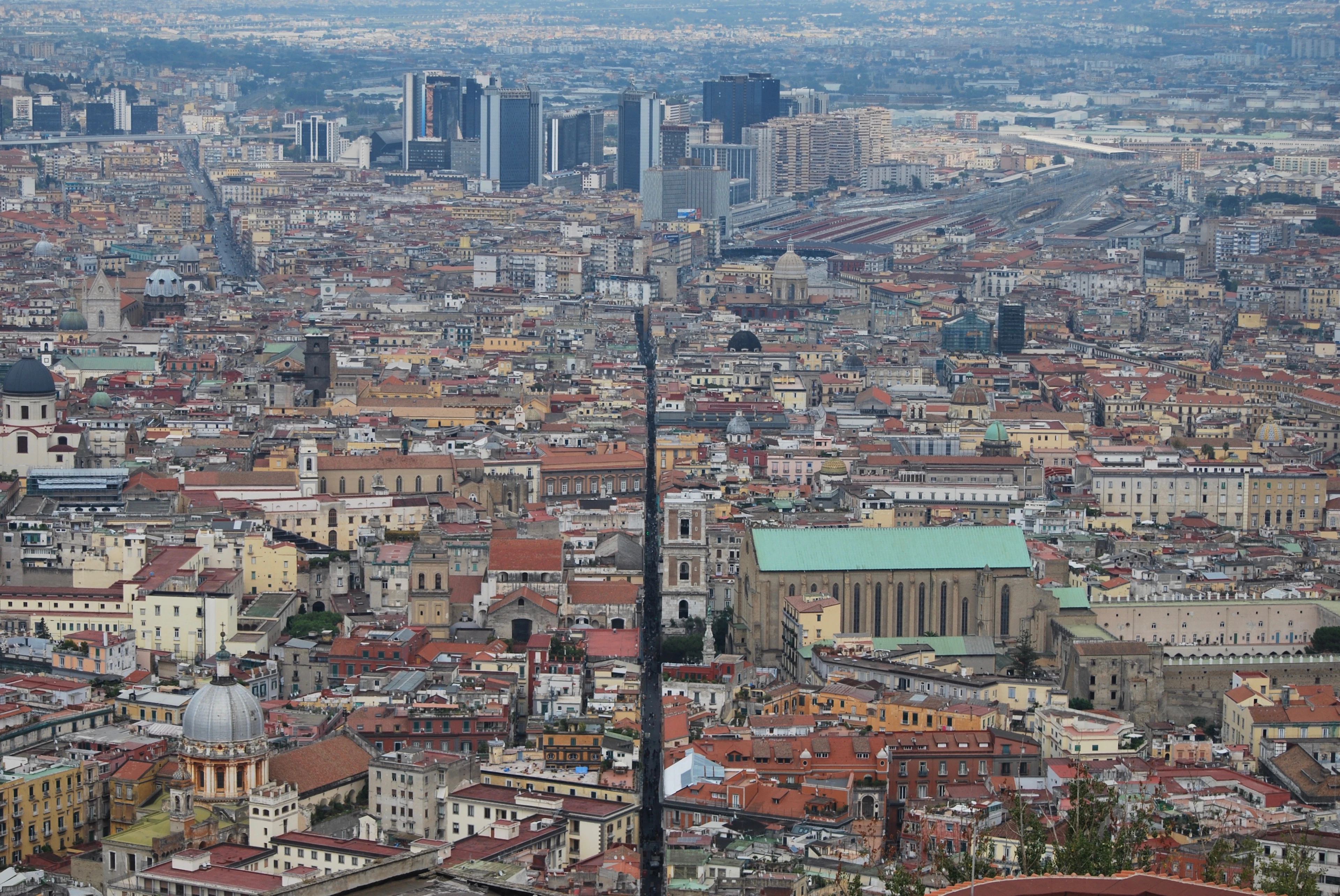 The centro storico of Naples seen from the hill above, a dense field of terracotta roofs, domes and bell towers with the arrow-straight lane of Spaccanapoli cutting through the middle of it.