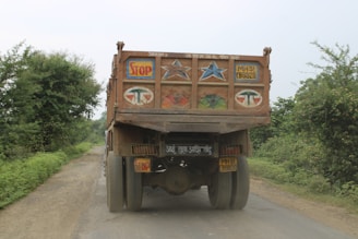 A heavily loaded truck is seen on a narrow rural road surrounded by greenery. The back of the truck features colorful decorations including stars and the word 'STOP'. The road is unpaved and appears dusty.