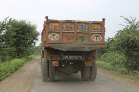 A heavily loaded truck is seen on a narrow rural road surrounded by greenery. The back of the truck features colorful decorations including stars and the word 'STOP'. The road is unpaved and appears dusty.