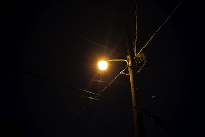 A streetlight illuminates a dark night, mounted on a wooden utility pole. The bright light creates a starburst effect with visible rays. Multiple electrical wires are attached to the pole, stretching out in different directions across the image.