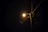 A streetlight illuminates a dark night, mounted on a wooden utility pole. The bright light creates a starburst effect with visible rays. Multiple electrical wires are attached to the pole, stretching out in different directions across the image.