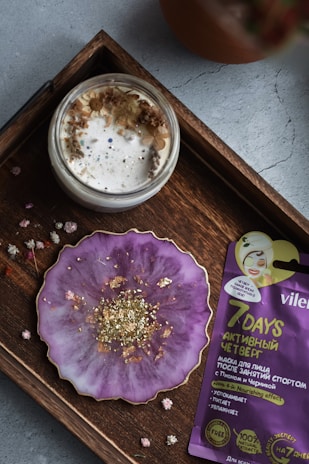 A wooden tray holds a small jar filled with cream topped with dried flowers and a purple, round plate with gold flakes. Beside the plate is a purple and yellow facial mask packet with text.