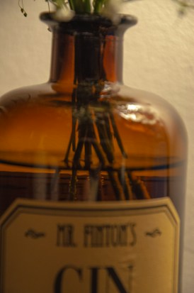 A brown glass bottle with a vintage label is partially filled with stems and blurred flowers extending from the top. The bottle reflects light, creating a warm glow against a soft, neutral background.