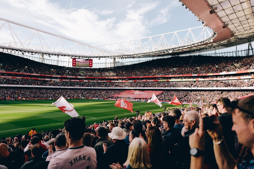 A large football stadium filled with spectators cheering and waving flags. The field is green and well-maintained, and the stands are packed with people. The atmosphere appears vibrant and energetic as people clap and celebrate.