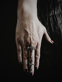 A hand with red painted nails is partially clenched, wearing a silver ring and holding a silver chain with a cross pendant. The background is dark, emphasizing the details of the hand and jewelry.