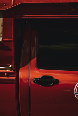 A close-up of a red vehicle door featuring a glossy finish and black door handle. The lighting creates dramatic shadows on the surface, highlighting the curves and contours. Part of a logo or emblem is partially visible on the bottom right.
