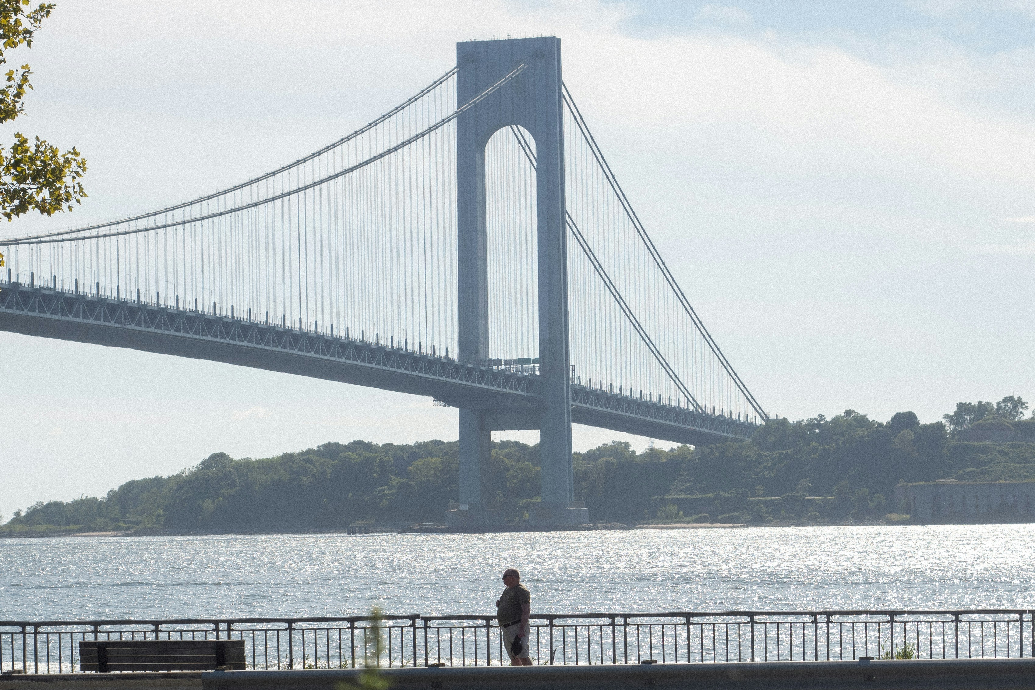 a person standing on the Shore Promenade, with the span and Staten Island tower  Verrazano–Narrows Bridge in the background.