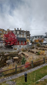 A picturesque scene featuring a charming two-story house with a red exterior and a dark roof, nestled among vibrant autumn foliage. A striking tree with bright red leaves stands prominently to the left, while neatly landscaped gardens with boulders and a small pond are visible in the foreground. In the background, large buildings with dormer windows and muted colors complete the tranquil setting.