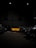 A dimly lit warehouse with a bright yellow sports car in the center, surrounded by other darker vehicles. The floor is cluttered with cardboard boxes and debris, while metallic shelves line the sides. The atmosphere appears industrial with sparse lighting overhead.