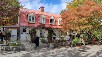 A quaint, rustic building with a red roof and beige siding, surrounded by autumnal trees. There is a sign for a crepe shop and outdoor seating with string lights above. The leaves on the trees display vibrant fall colors, creating a cozy atmosphere.