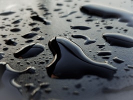 Large and small droplets of a dark liquid, possibly oil, are scattered on a smooth, reflective surface. The droplets vary in size and shape, with some merging together. The surface appears wet and shiny, reflecting light subtly.
