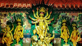 A vibrant and ornate depiction of a Hindu goddess with multiple arms stands at the center, surrounded by other deities, including an elephant-headed figure. The background is adorned with colorful tribal-style art, and the scene is framed with rich and intricate patterns.