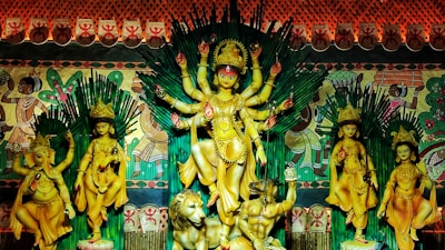 A vibrant and ornate depiction of a Hindu goddess with multiple arms stands at the center, surrounded by other deities, including an elephant-headed figure. The background is adorned with colorful tribal-style art, and the scene is framed with rich and intricate patterns.