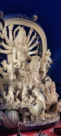 A detailed sculpture of a multi-armed deity seated on a majestic lion, surrounded by smaller figures. The sculpture is intricate, featuring ornate jewelry and divine symbols in a light, possibly made of plaster or clay. The background is dark blue, enhancing the focus on the sculpture.