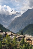 A serene village nestled in a valley, surrounded by lush green trees and towering mountains. The peaks of the mountains are capped with snow and partially obscured by clouds, providing a dramatic backdrop. The rooftops of the village houses are clearly visible, blending harmoniously with the natural surroundings.