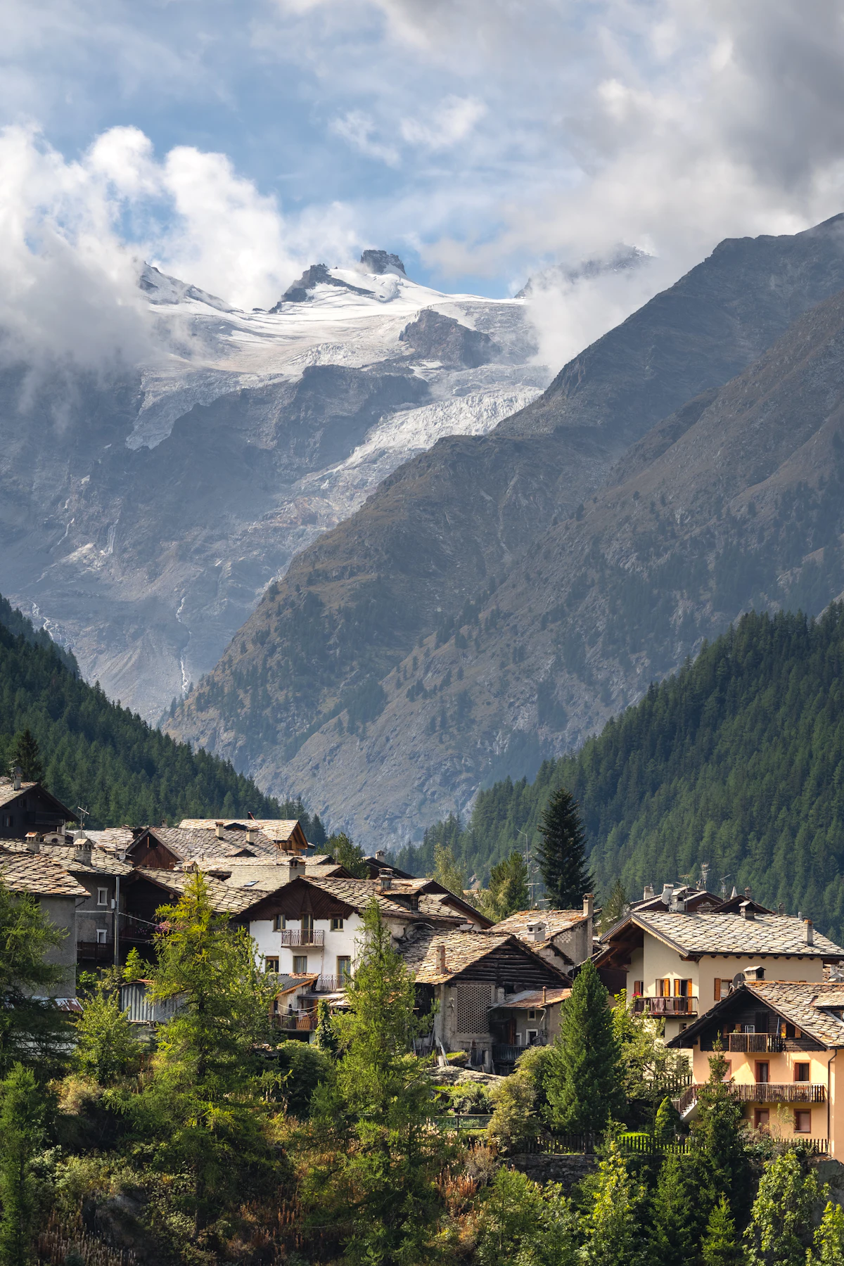 Houses and conifers in Valsavarenche, with the glaciers of Gran Paradiso in the clouds behind
