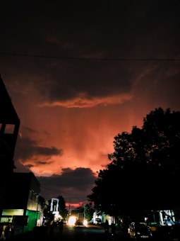 A city street at dusk with a dramatic, dark sky illuminated by a reddish-orange glow from the setting sun, casting shadows on the buildings and trees. Illuminating headlights of a vehicle in the background and silhouettes of trees and structures add to the atmospheric scene.