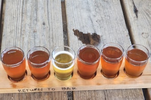 A wooden flight board holds six small glasses, each filled with different shades of beer, ranging from amber to pale yellow. The board is set on a rustic wooden table and features a handwritten note 'RETURN TO BAR' in black marker.