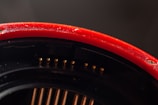 A close-up view of the interior of a mechanical or electronic device, featuring a smooth, glossy red exterior curved surface, accented by a series of small, metallic gold pins arranged linearly along the black part of the component.