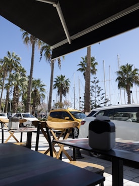 An outdoor seating area with wooden chairs and tables under a black canopy. Palm trees line the background, alongside parked cars including a yellow taxi. Sailboat masts are visible beyond the trees, suggesting a nearby marina.