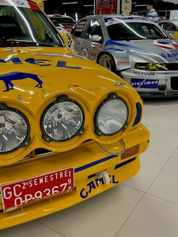 A close-up view of a vintage yellow race car with prominent rally headlights and a red license plate. The car's hood features branding and artwork, and it is parked in an indoor showroom alongside other racing cars. Another car seen in the background has a silver and blue color scheme with various logos.