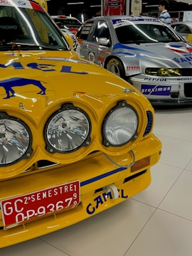 A close-up view of a vintage yellow race car with prominent rally headlights and a red license plate. The car's hood features branding and artwork, and it is parked in an indoor showroom alongside other racing cars. Another car seen in the background has a silver and blue color scheme with various logos.